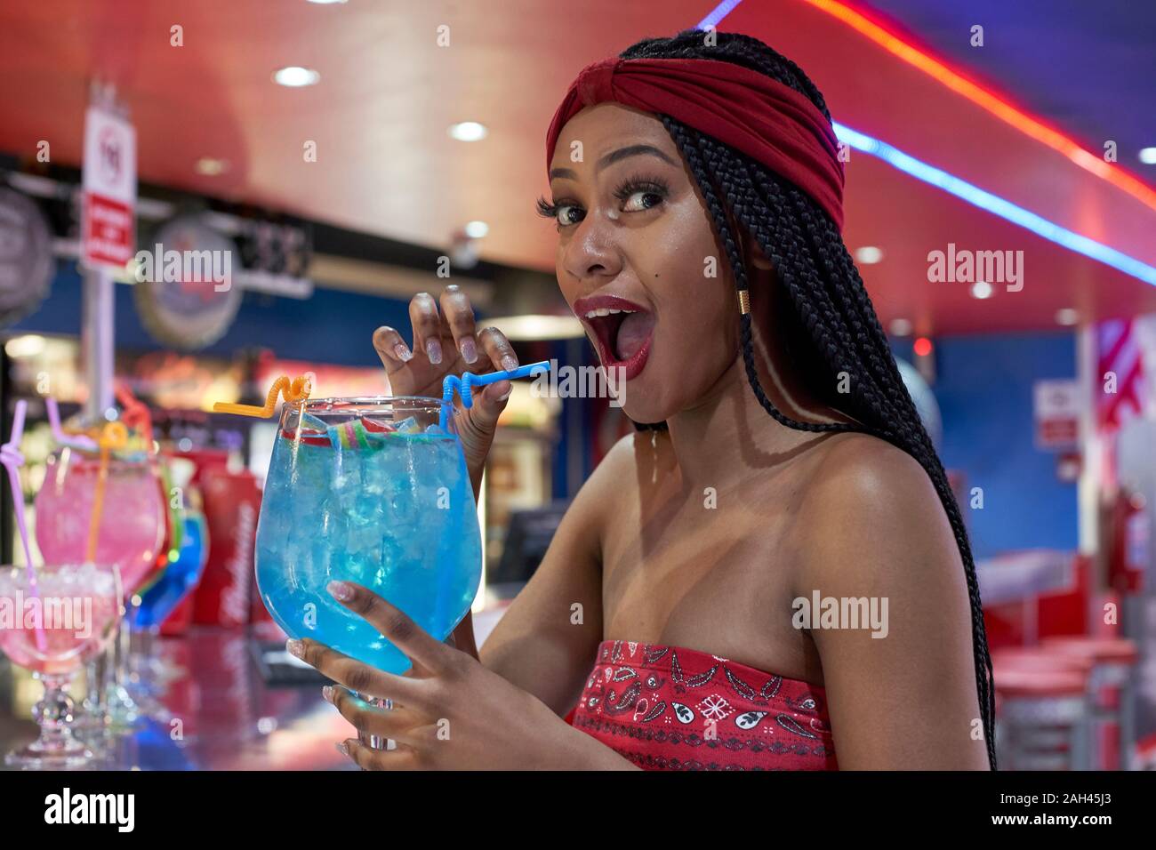 Young woman with braided hairstyle sitting on a restaurant's bar, drinking a blue cocktail Stock Photo