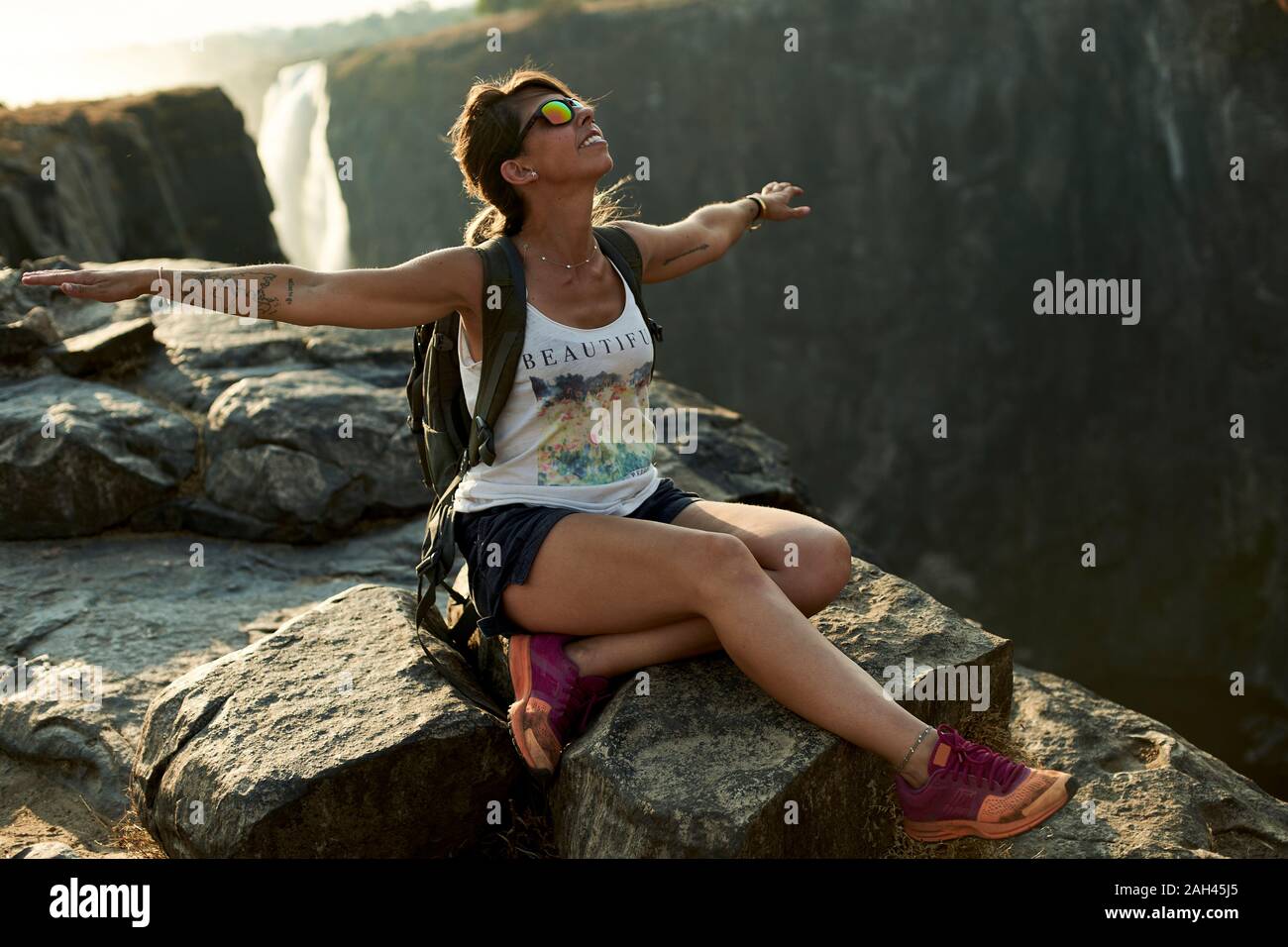 Happy woman sitting with arms open on top of a rock at Victoria Falls at sunset, Zimbabwe Stock Photo