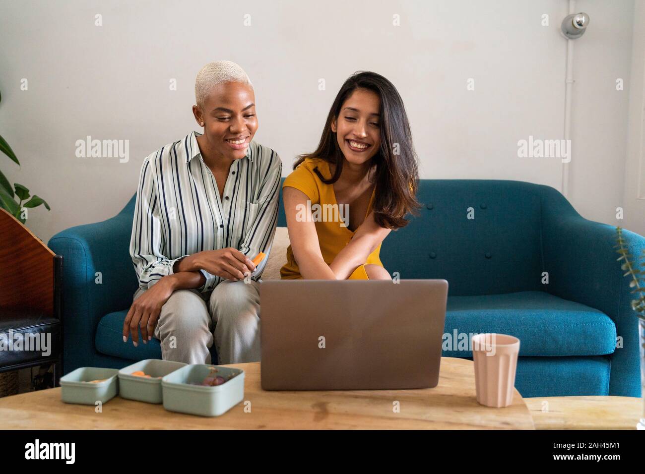 Two happy women sitting on couch using laptop together Stock Photo