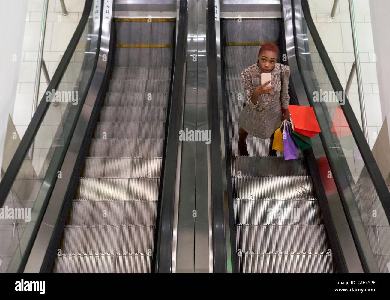 Young woman with holding colorful shopping bags and using her phone while standing on escalator Stock Photo