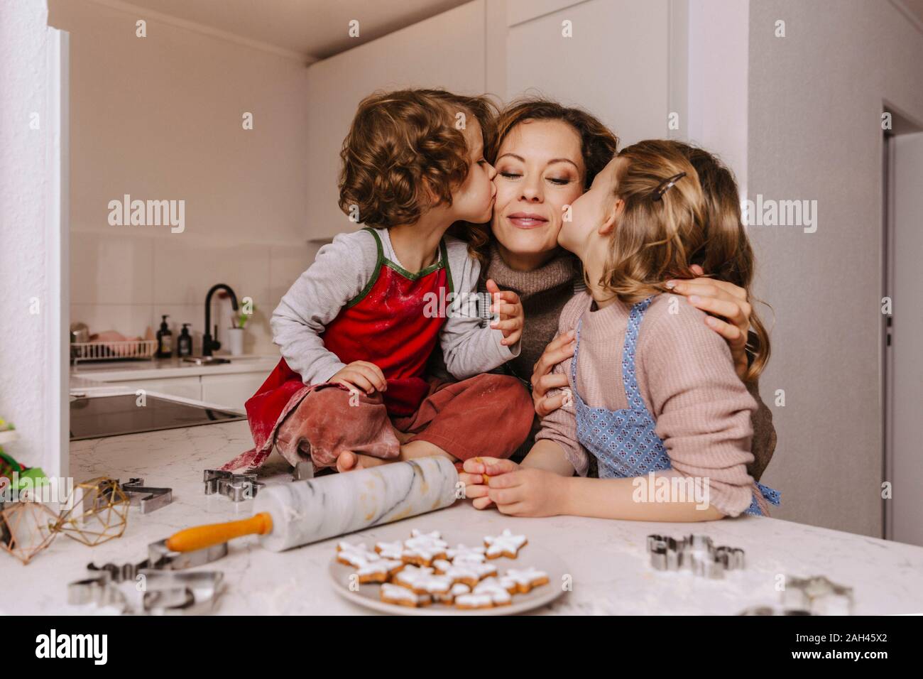 Girls kissing mother in kitchen with Christmas cookies on counter Stock Photo