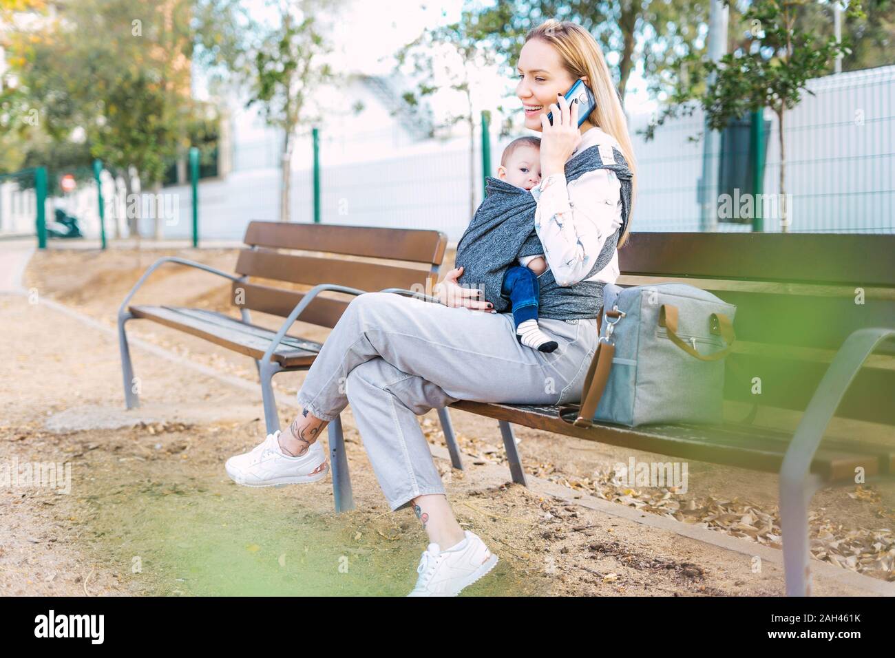 Happy mother resting with baby boy on a park bench talking on the phone ...