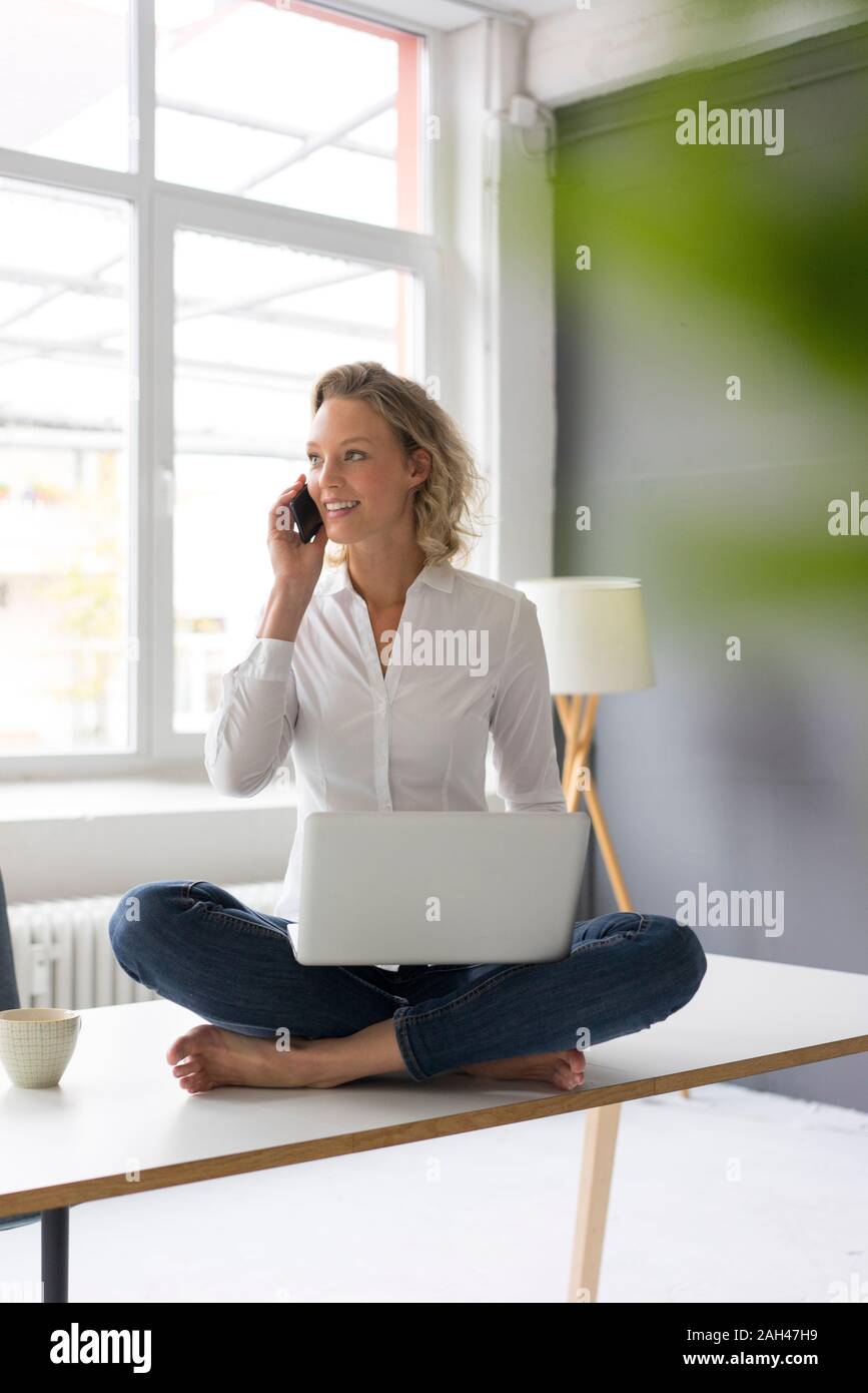 Smiling young businesswoman sitting on desk in office using laptop and cell phone Stock Photo