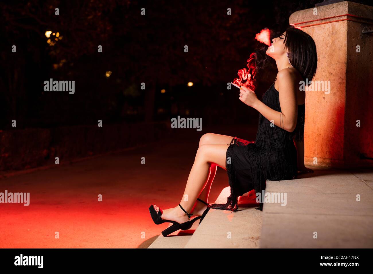 Smoking young woman wearing black evening dress sitting on stairs at night Stock Photo