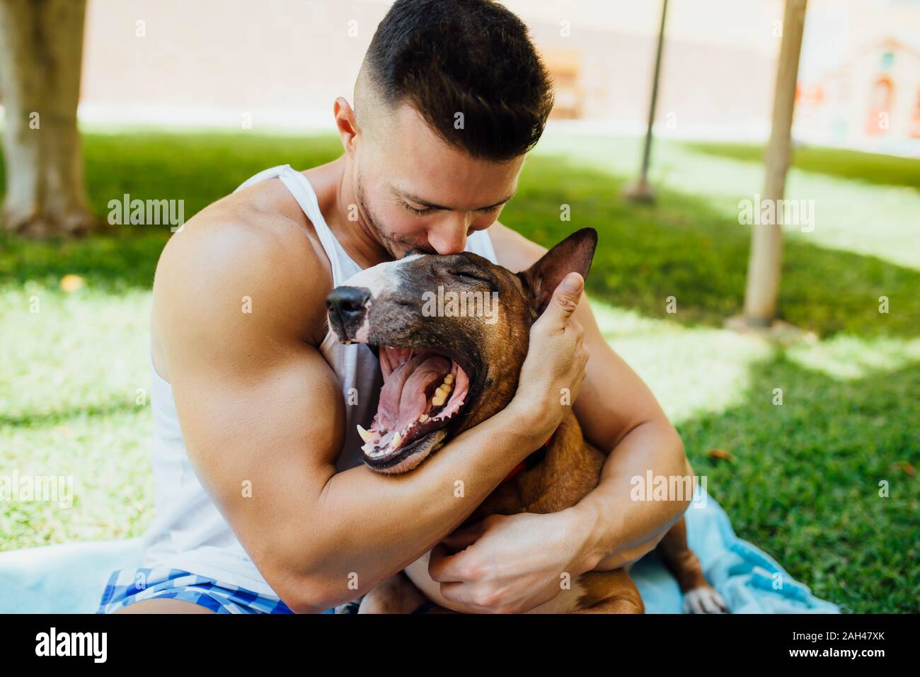 Muscular man sitting on blanket on a meadow hugging his dog Stock Photo