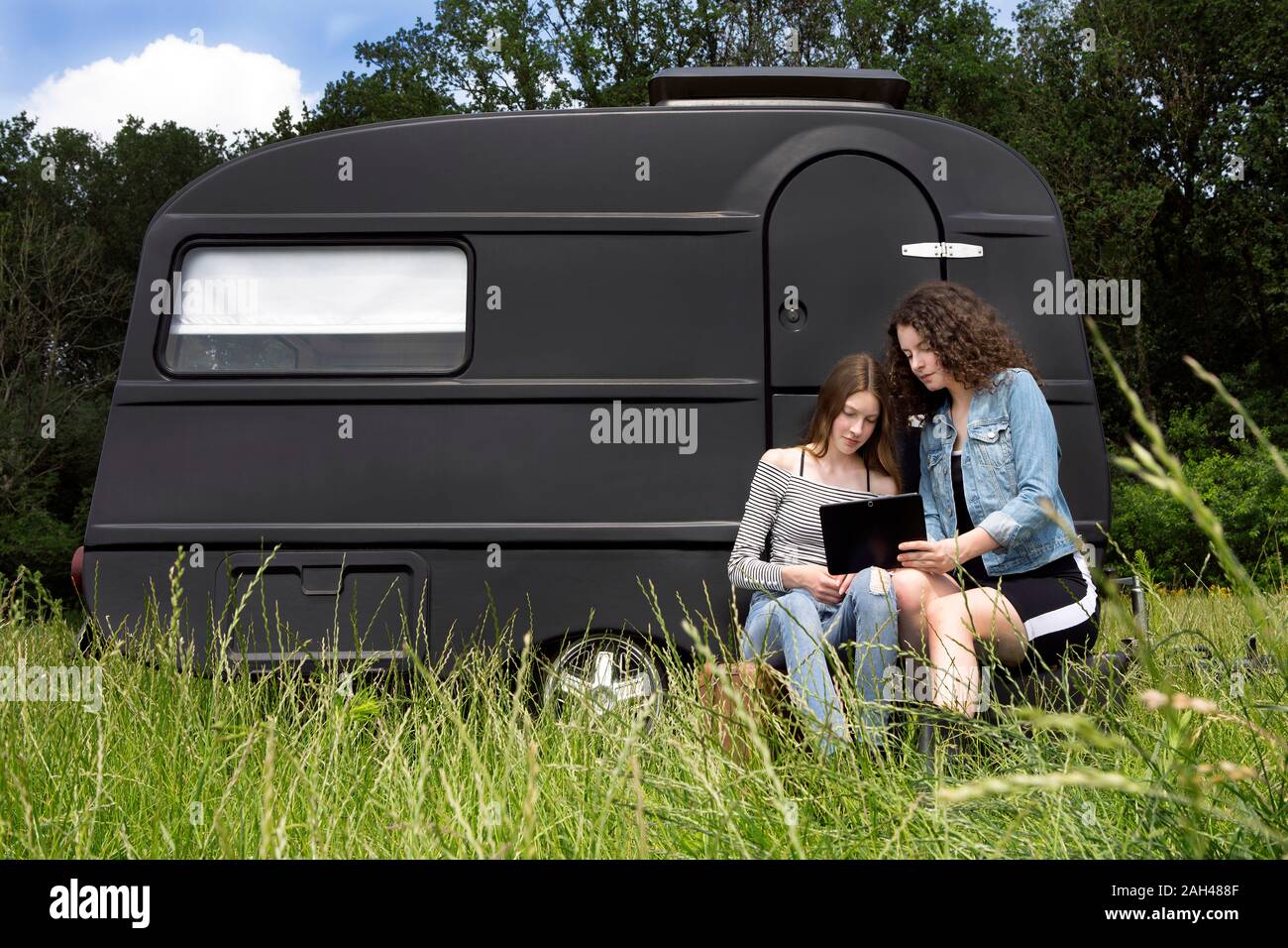 Two friends sitting on a meadow in front of black caravan using digital tablet Stock Photo