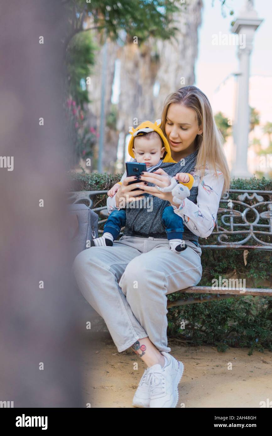 Mother resting with baby boy on a park bench using smartphone Stock ...