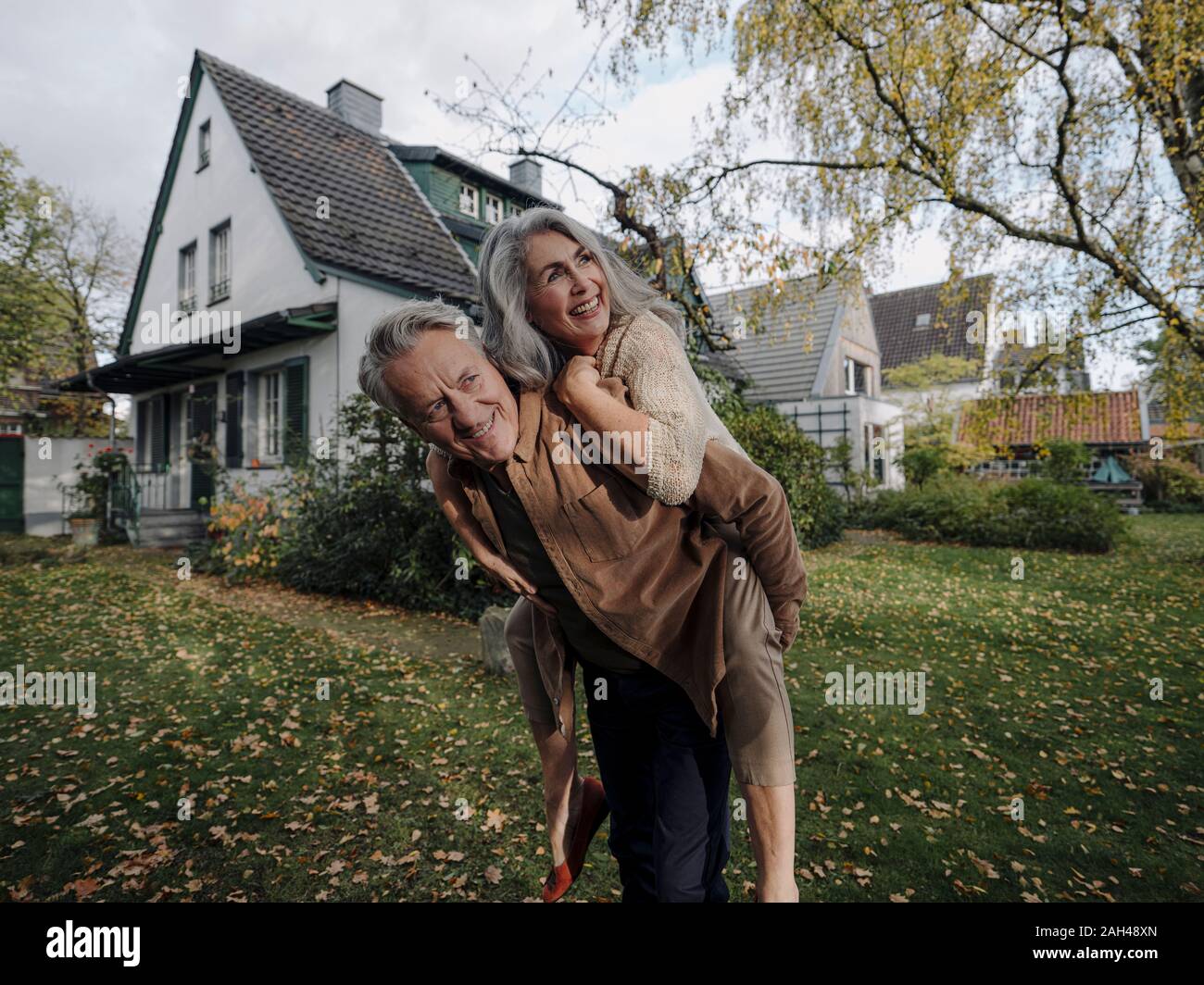 Happy senior man carrying wife piggyback in garden of their home in autumn Stock Photo