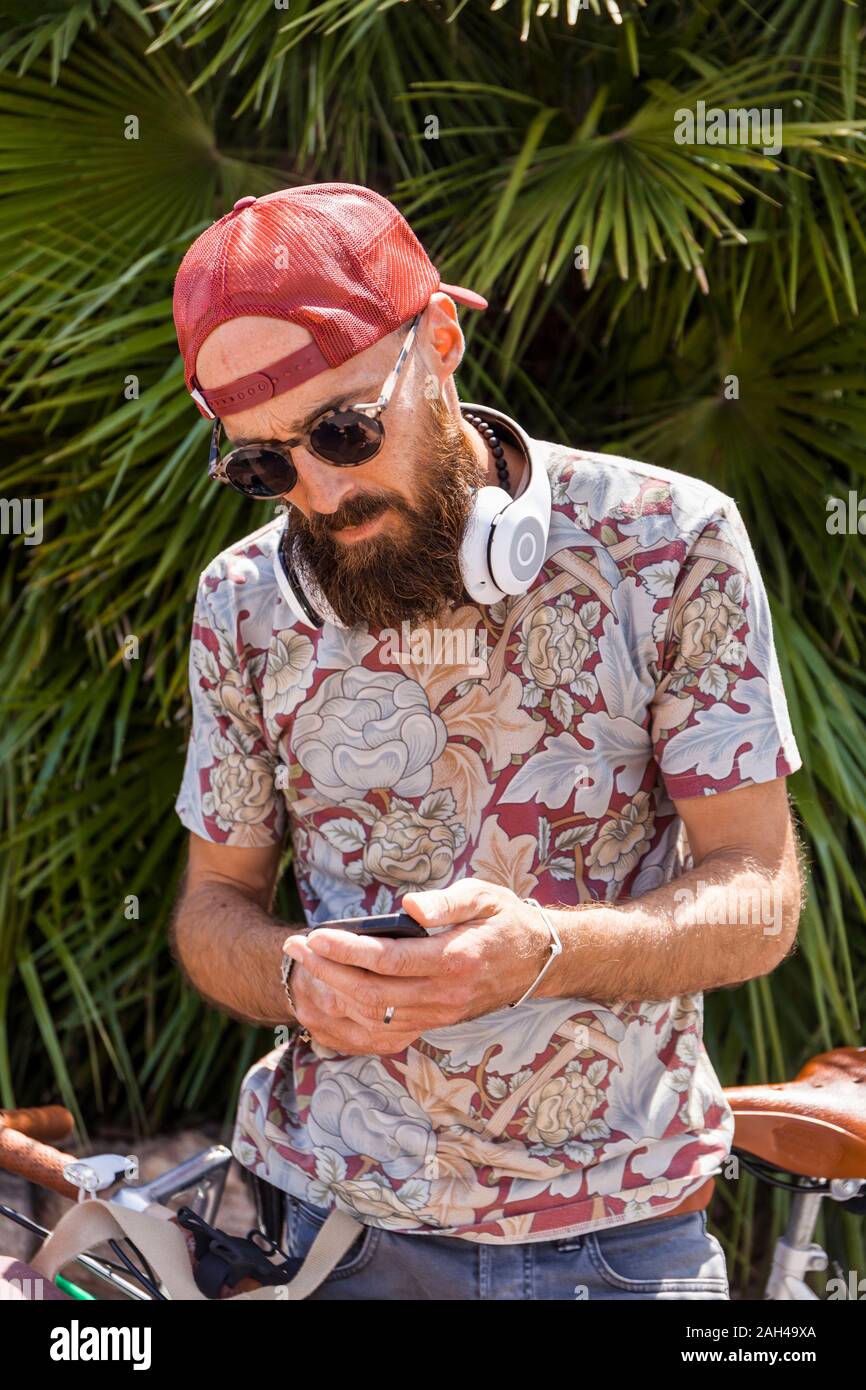 Mature man with red basecap, sunglasses and white headphones using smartphone Stock Photo