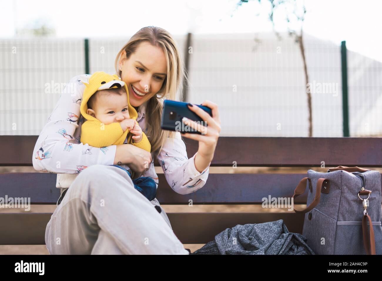 Happy mother resting with baby boy on a park bench taking a selfie ...