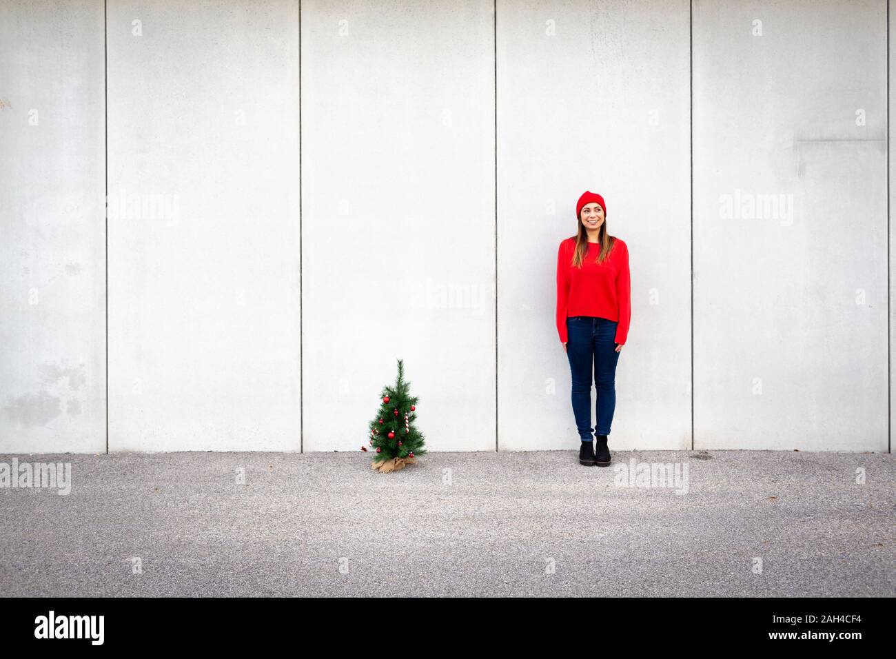 Woman wearing red pullover and wolly hat, holding artificial Christmas tree in front of a wall Stock Photo