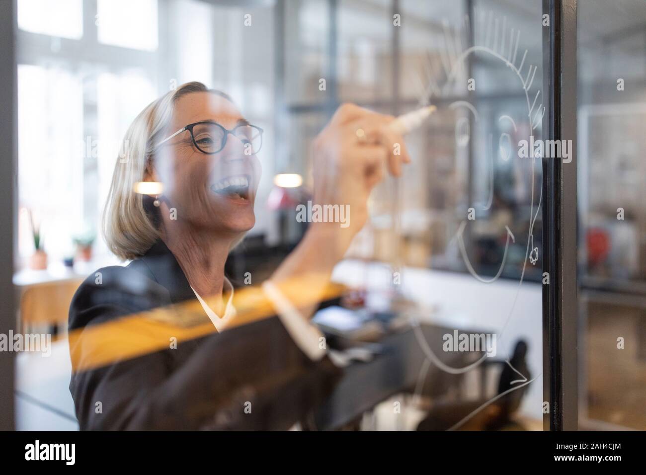 Happy mature businesswoman drawing face on glass pane in office Stock Photo