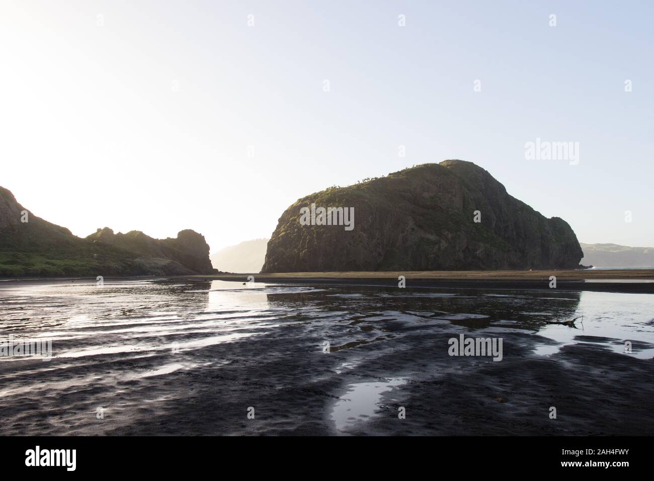 Early morning at the black sand beach Whatipu in the Waitakere Ranges of West Auckland, New Zealand. There are steep rock mountains reflected in water. Stock Photo