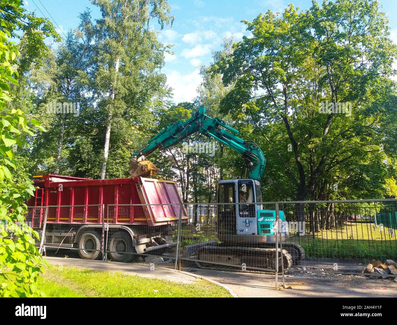 Road repair. In the courtyard of the city, amid grass and trees behind a metal fence, a turquoise excavator digs up a roadbed. Nearby is a red dump truck. Russia, the city of St. Petersburg, August 2019. Stock Photo