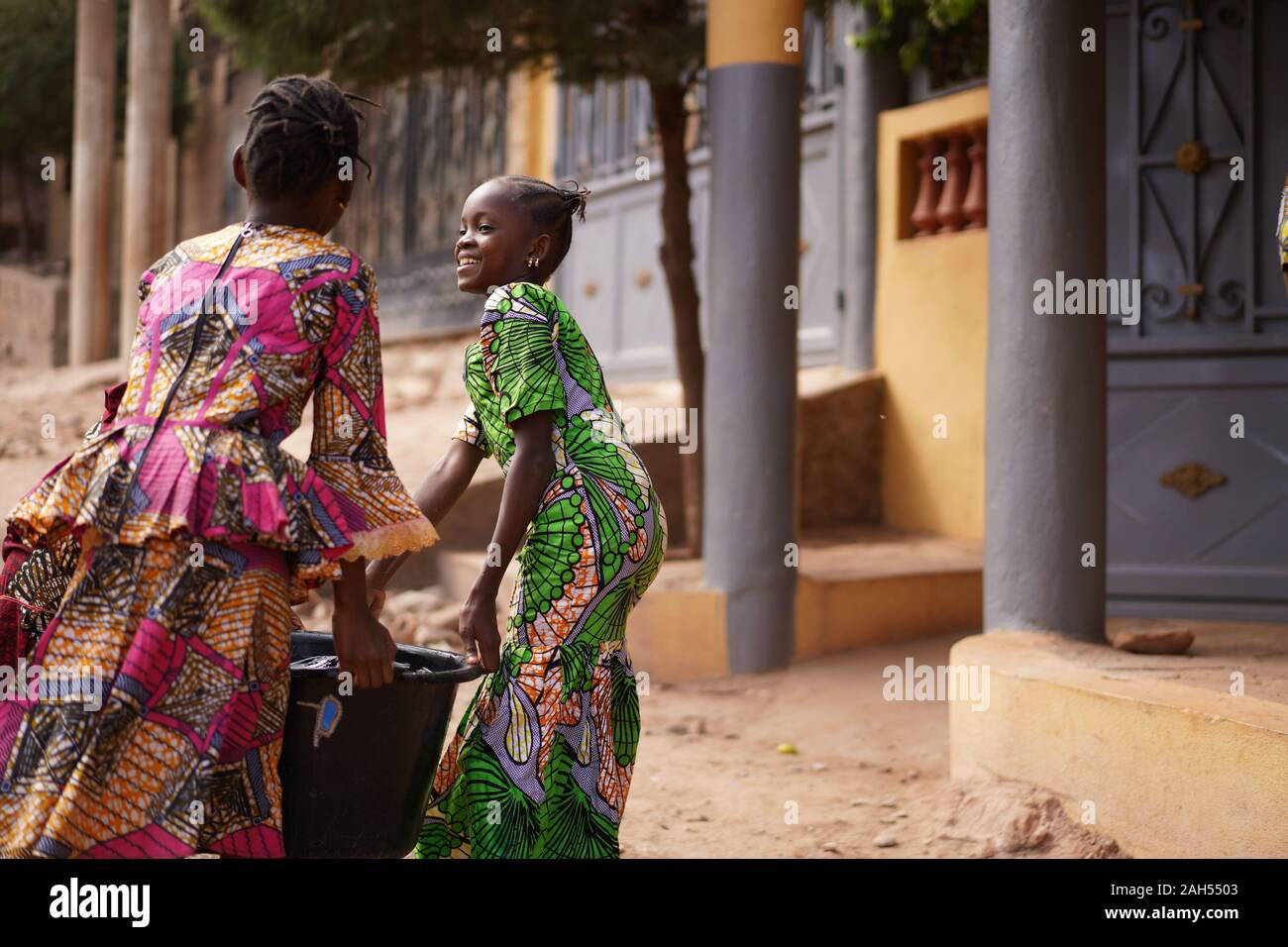 Two African Girls Carrying A Heavy Water Bucket Home From The Pumping Station Stock Photo