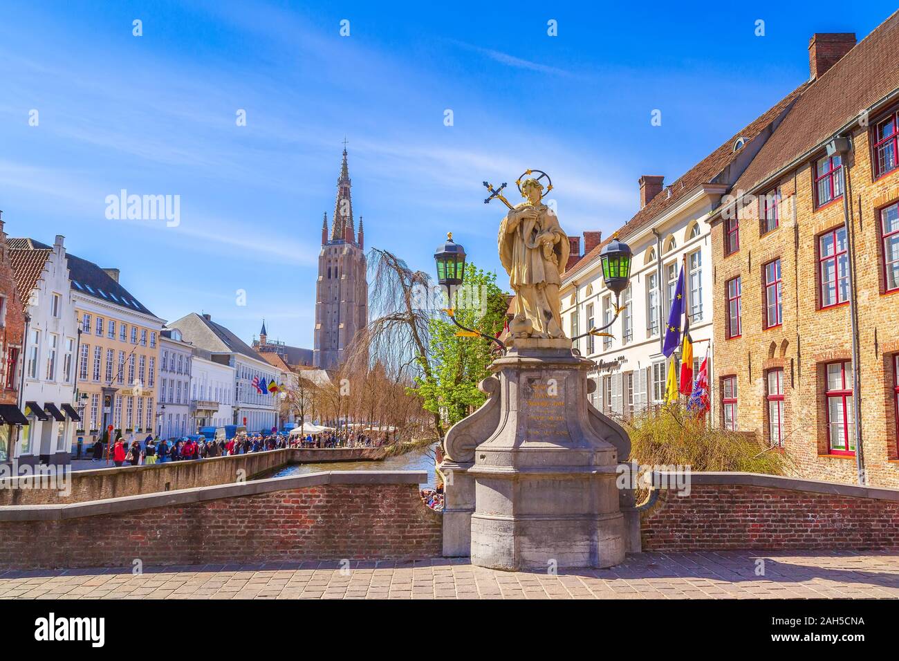Bruges, Belgium - April 10, 2016: Street and canal view with houses, statue of Johannes Nepomucenus, church tower and people in Brugge Stock Photo