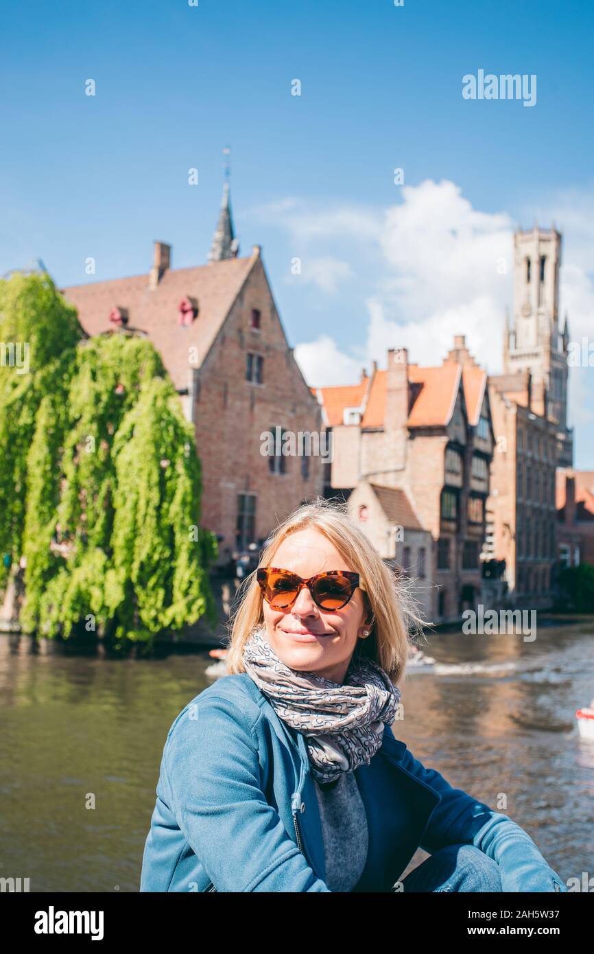 A beautiful young girl in casual clothes sits on the background of a famous tourist spot with a canal in the historical center of Bruges, Belgium Stock Photo