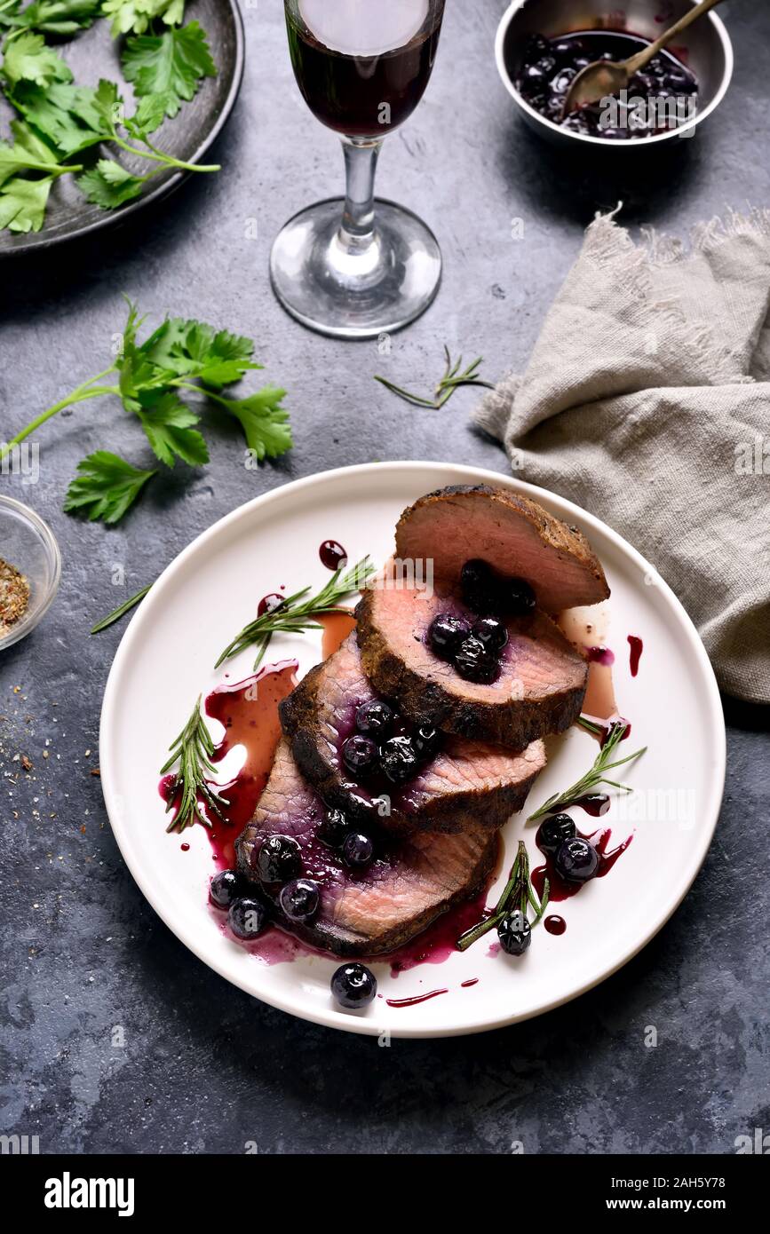 Sliced grilled beef with blueberry sauce on white plate over blue stone background. Tasty medium rare roast beef with berry sauce. Stock Photo