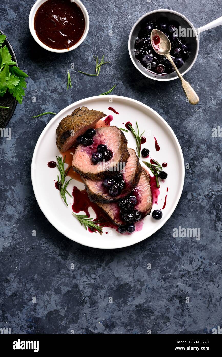 Sliced grilled beef with blueberry sauce on white plate over blue stone background with free space. Tasty medium rare roast beef with berry sauce. Top Stock Photo