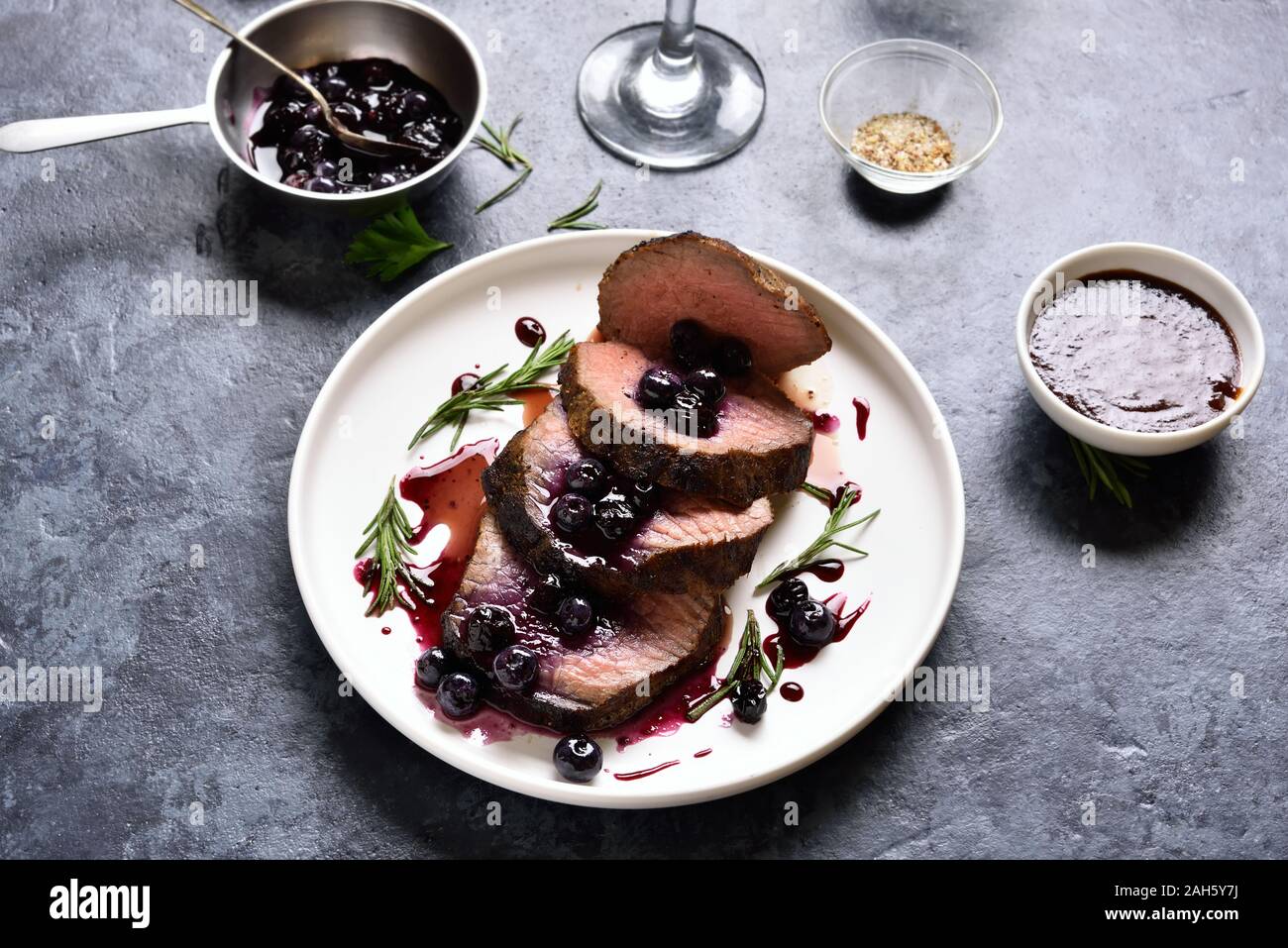 Sliced grilled beef with blueberry sauce on white plate over dark stone background. Tasty medium rare roast beef with berry sauce. Stock Photo