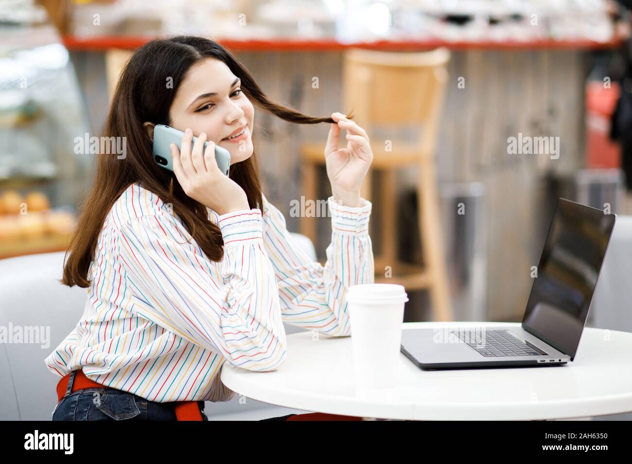 Pretty young brunette girl sitting in cafe and working on laptop, talking on phone, drinking coffee Stock Photo