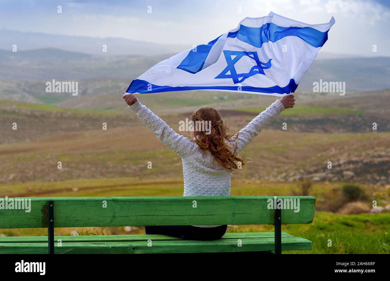 Beautiful young jewish girl sitting holding Israel flag in the wind and enjoying great view landscape on the sky, field and mountains.Patriotic holiday. Independence day Israel - Yom Ha'atzmaut . Stock Photo