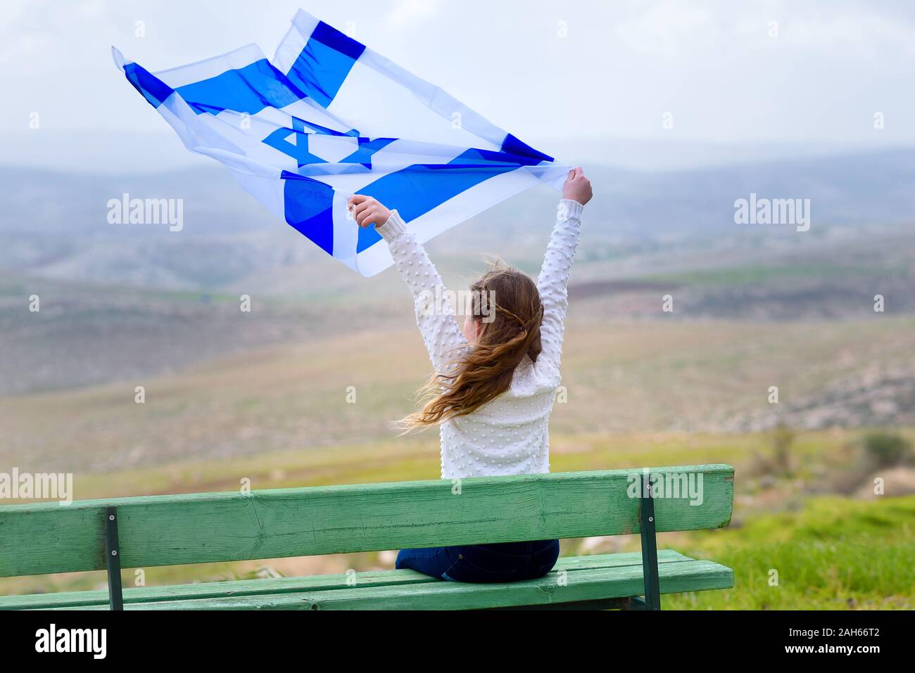Beautiful young jewish girl sitting holding Israel flag in the wind and enjoying great view landscape on the sky, field and mountains.Patriotic holiday. Independence day Israel - Yom Ha'atzmaut . Stock Photo