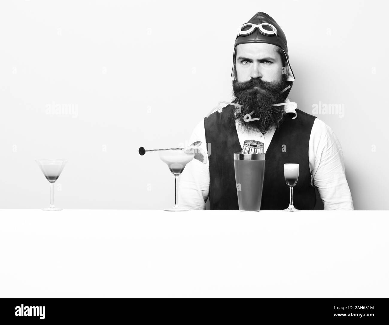 handsome bearded aviator or pilot with long beard and mustache on serious face with shaker alcoholic cocktail in vintage suede leather, waistcoat with hand and glasses on purple green studio background Stock Photo