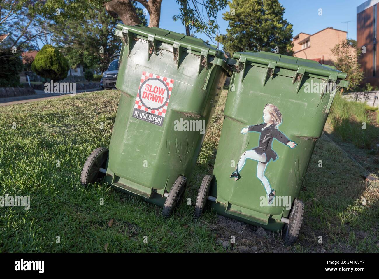Two large 240L green waste bins with road safety concept stickers or decals attached sit on a roadside footpath facing the morning sun in Sydney, Aust Stock Photo