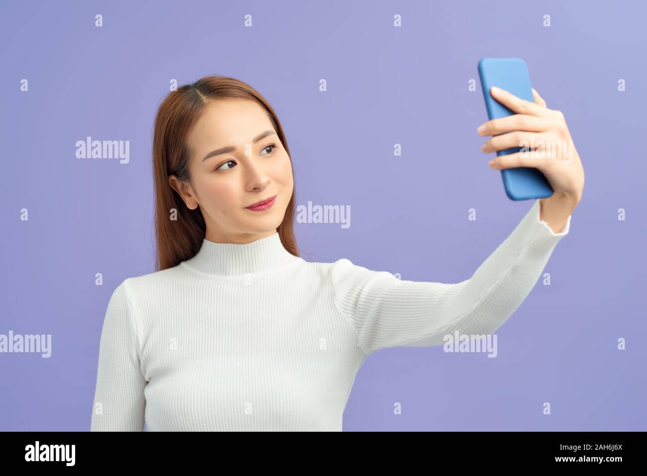 Selfie time! Attractive young woman in casual wear making selfie by her smart phone and smiling while standing against violet background Stock Photo