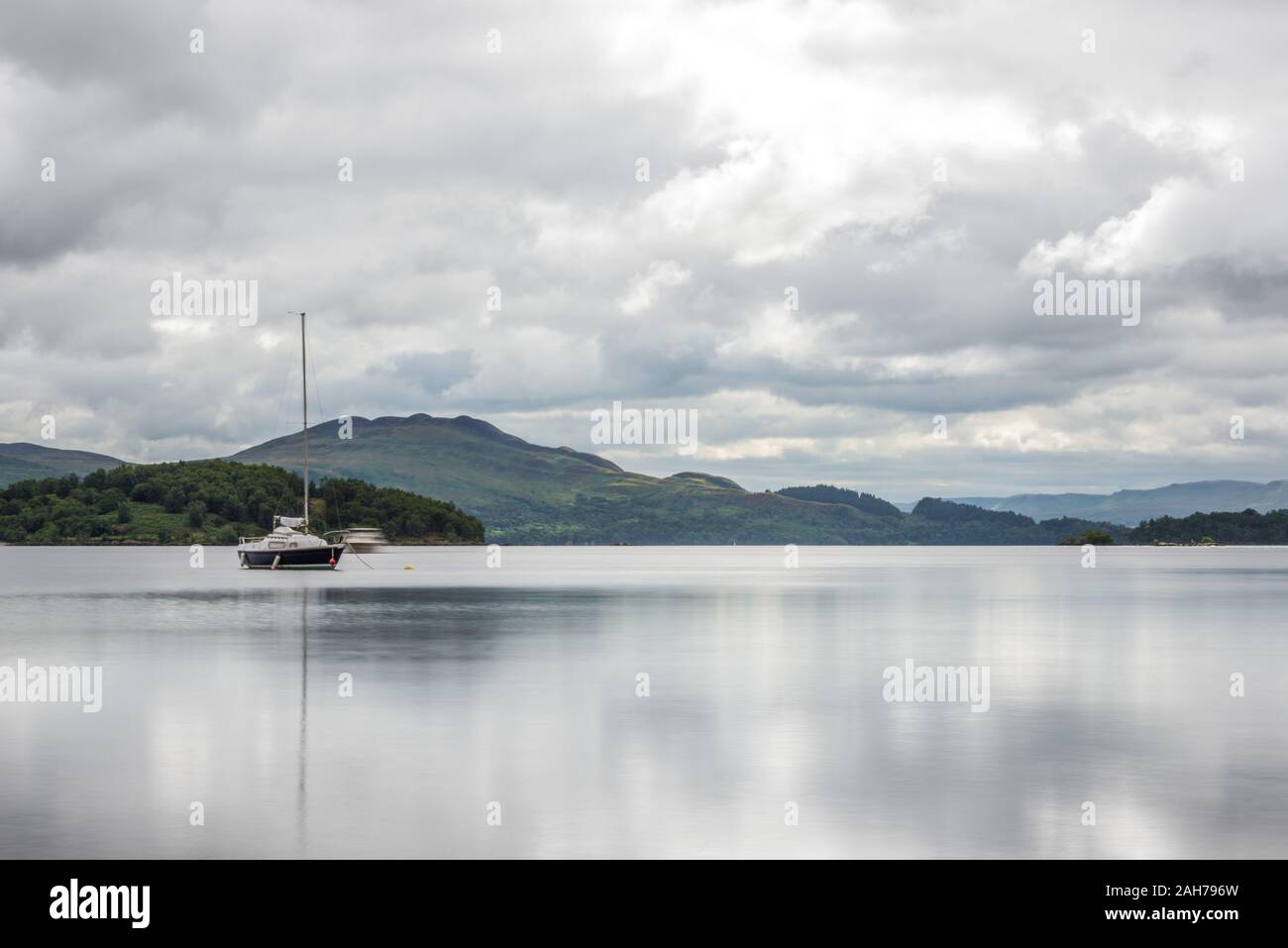 A scottish lake surrounded by trees and vegetation with a sailboat in the middle and distant mountains in the background, under a cloudy sky Stock Photo