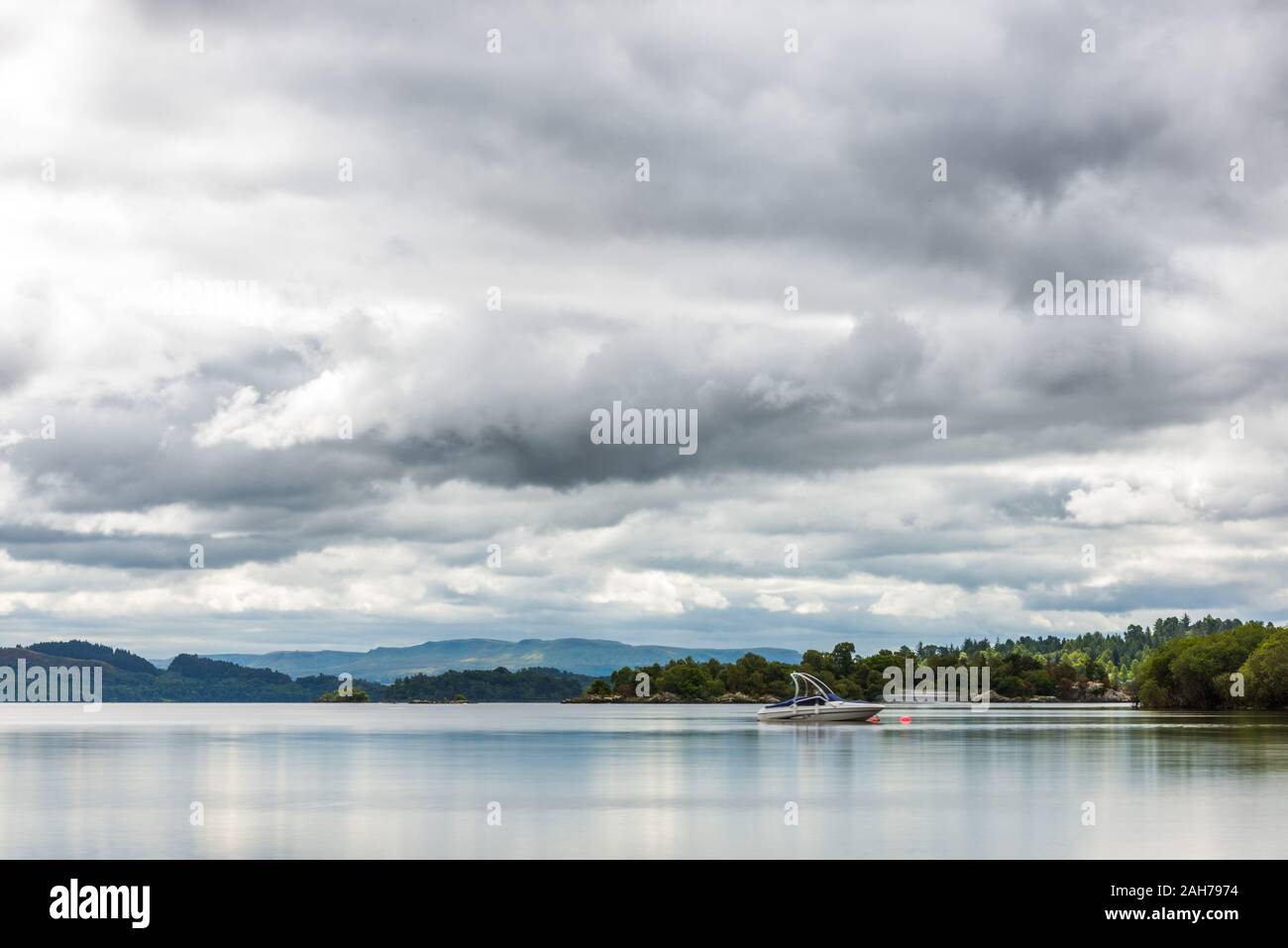 A scottish lake surrounded by trees and vegetation, with a motorboat moored in the middle and distant mountains under a cloudy sky Stock Photo