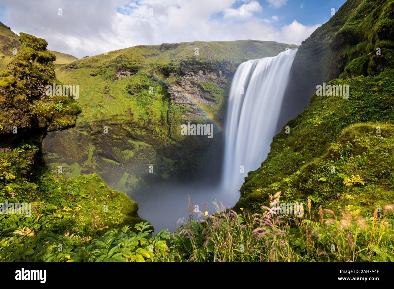 Long exposure of the icelandic waterfall of Skogafoss, surrounded by cliffs covered with green musk, under a blue sky with clouds Stock Photo