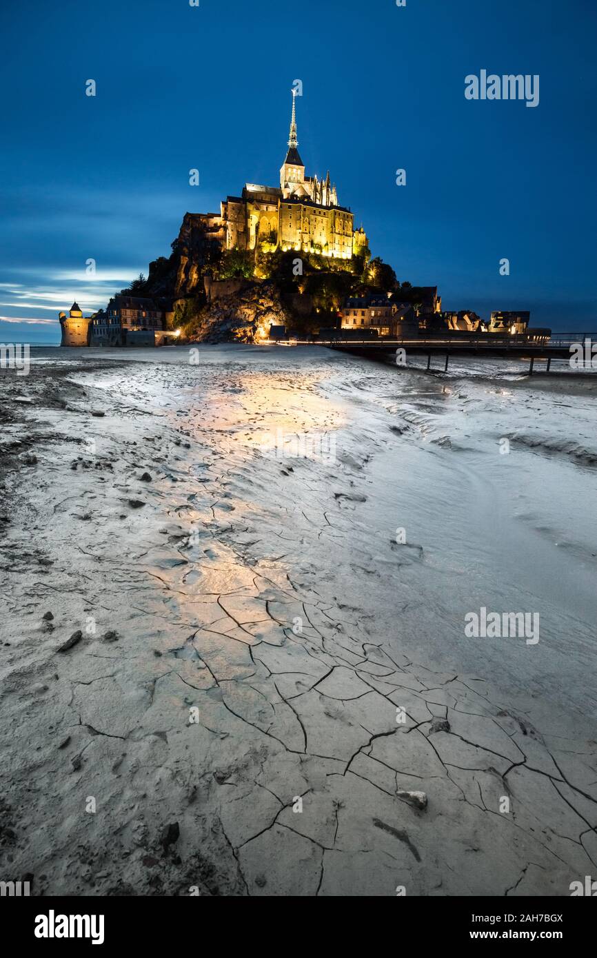 Night shot of the french stronghold of Le Mont-Saint-Michel, with its lights reflecting on the cracked gray clay in the foreground, against a blue sky Stock Photo