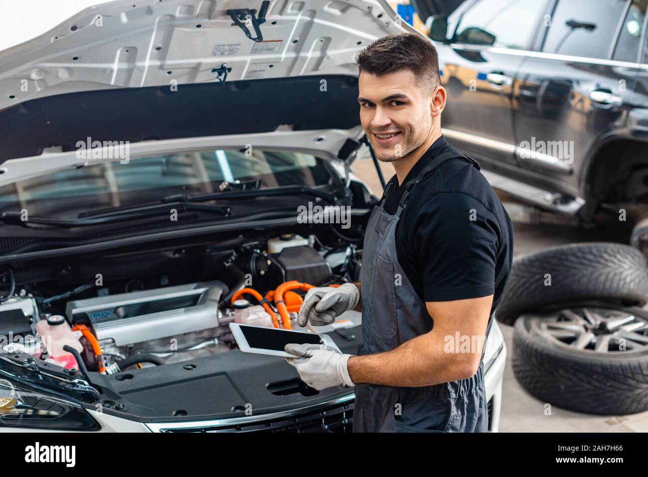 handsome mechanic smiling at camera while using digital tablet near car engine compartment Stock Photo