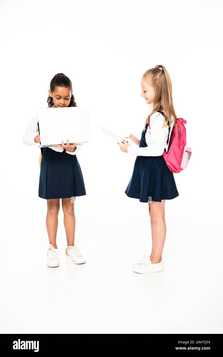 full length view of two multicultural schoolgirls using laptop and digital tablet on white background Stock Photo