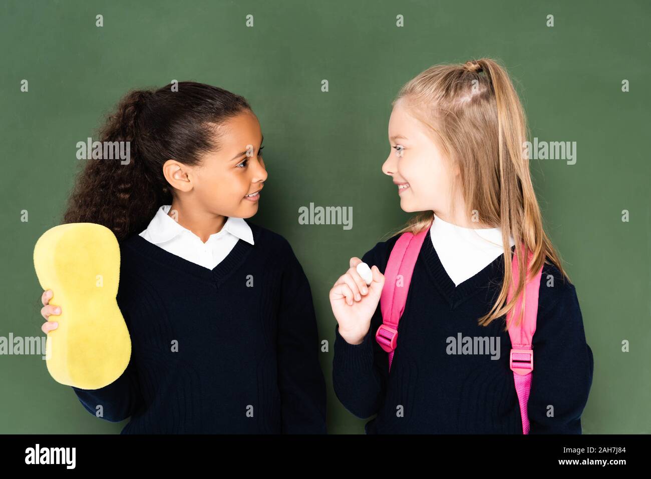 two cheerful multicultural schoolgirls looking to each other while standing near chalkboard Stock Photo