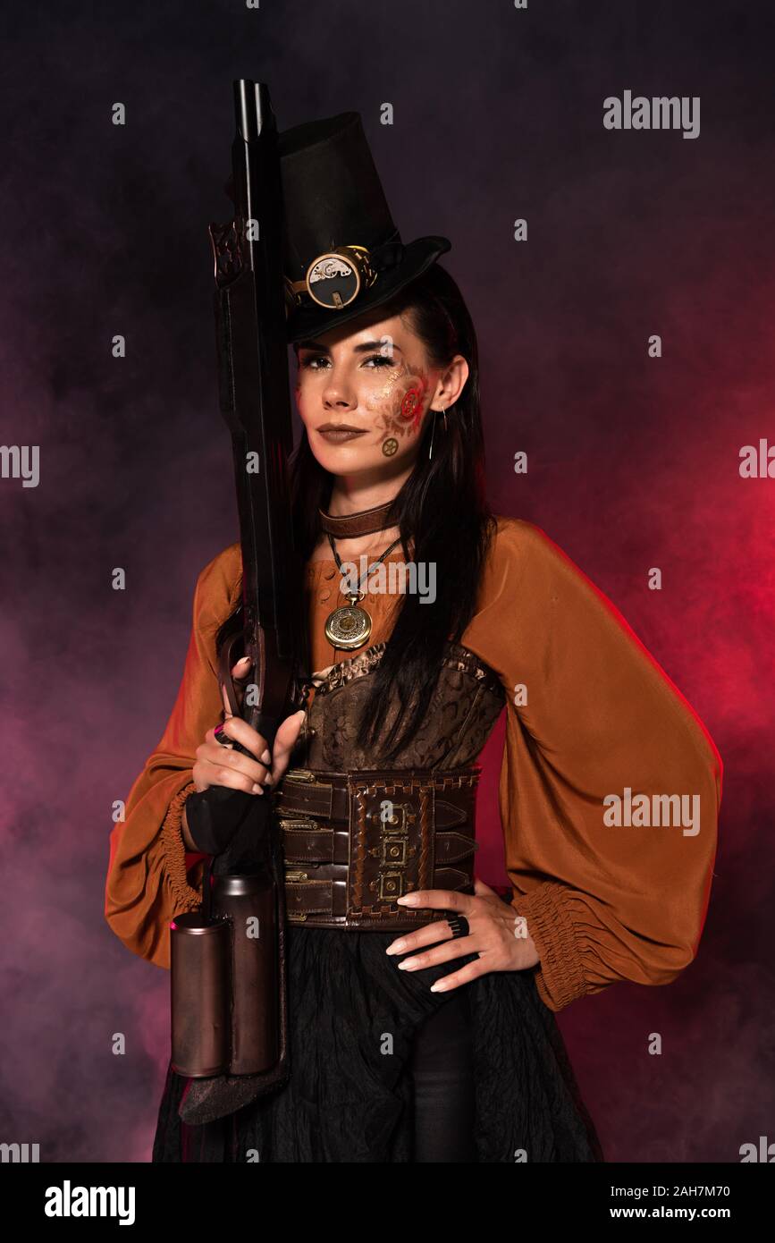 attractive steampunk woman holding gun and standing with hand on hip in pink smoke Stock Photo