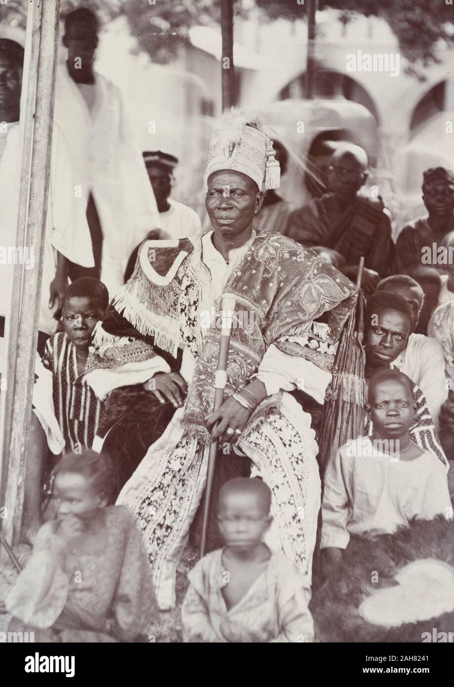 Nigeria, Marking on reverse: 'Badagry 1927'An unidentified chief, perhaps the Akran of Badagry, is seated wearing elaborate embroidered robes with a high-sided and tasselled cap. He holds a staff of office and is surrounded by children sitting on the ground, one of whom holds a fan edged with feathers, 1927. 2000/098/2/97. Stock Photo