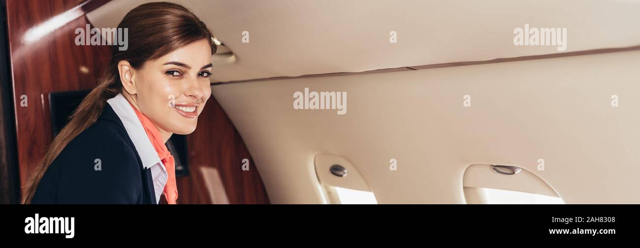 panoramic shot of smiling flight attendant in uniform looking at camera in private plane Stock Photo