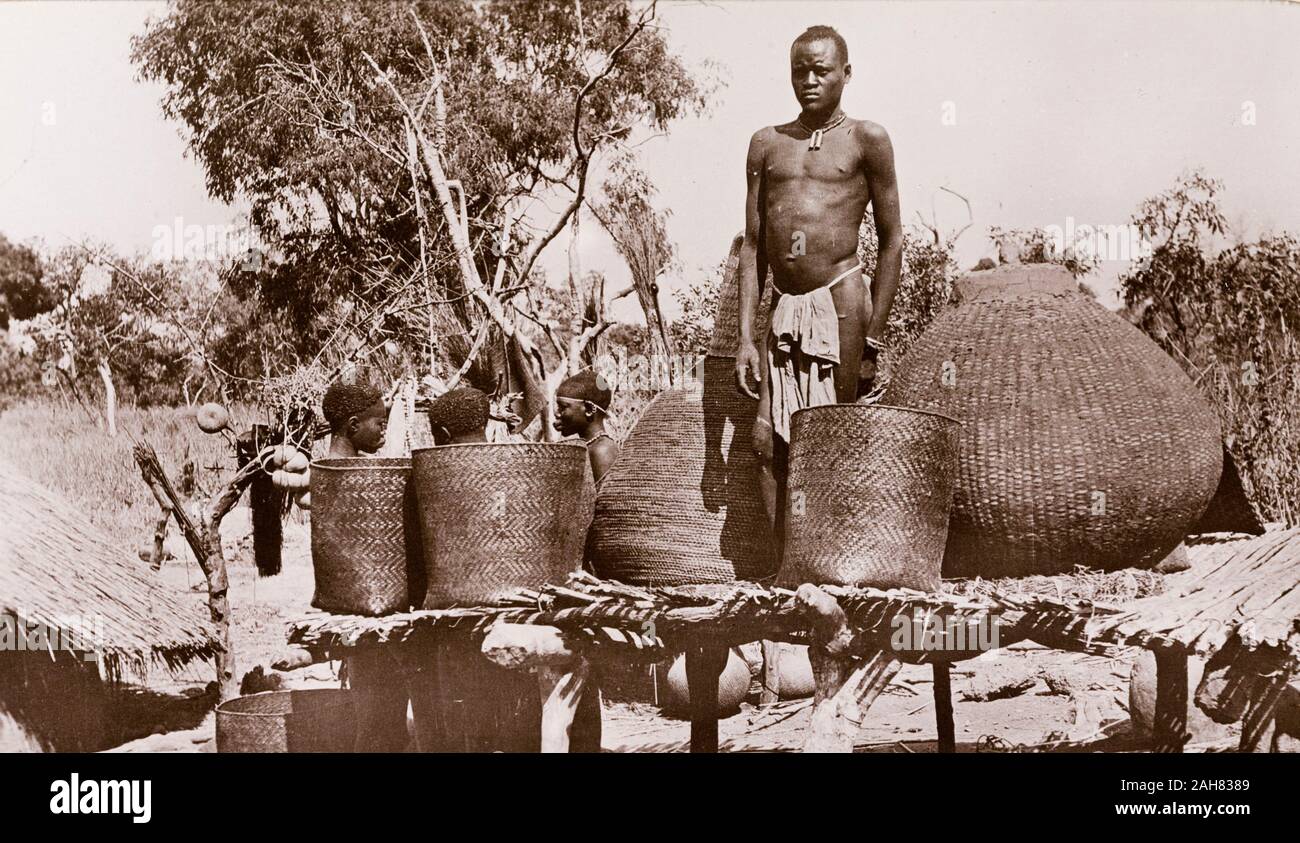 Sudan, Printed caption reads: 'Jurs Granary, Bahr-el-Ghazal. Published by G N Morhig, The English Pharmacy, Khartoum. Copyright 169'. A man in a loincloth stands on a raised platform between several baskets at a village granary. Three women chat behind him at ground level, [c.1906]. 2003/222/1/2/73. Stock Photo