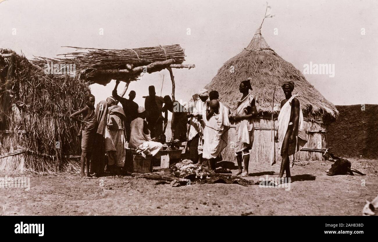 Sudan, Printed caption reads: 'A Butcher's Shop, Upper Nile. Published by G N Morhig, The English Pharmacy, Khartoum. Copyright 107'. A line of men queue up outside a village butcher's stall, waiting to be served by an assistant who sits at a workbench beneath a canopy, surrounded by cuts of meat, [c.1906]. 2003/222/1/2/79. Stock Photo