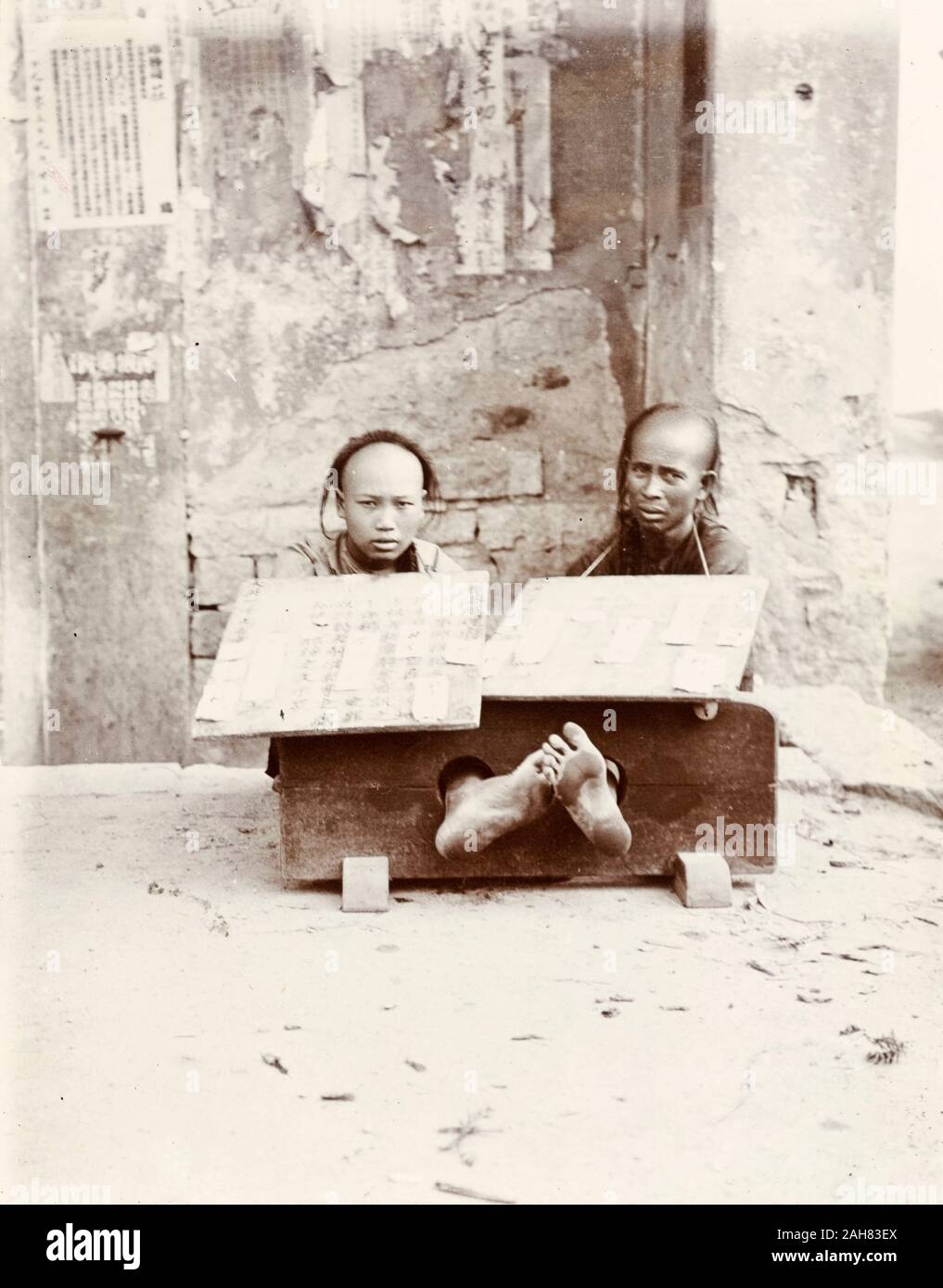 Hong KongChina, Two petty criminals sit with their ankles restrained in stocks, suffering public humiliation on a city street. Each wears a board strung around his neck, describing in detail the offense committed. Original manuscript caption: Prisoners in stocks. - generally for six hours at a stretch. Very uncomfortable if feet higher than level of ground sat upon. Their misdemeanor depicted on board, circa 1903. 1998/028/1/1/34. Stock Photo