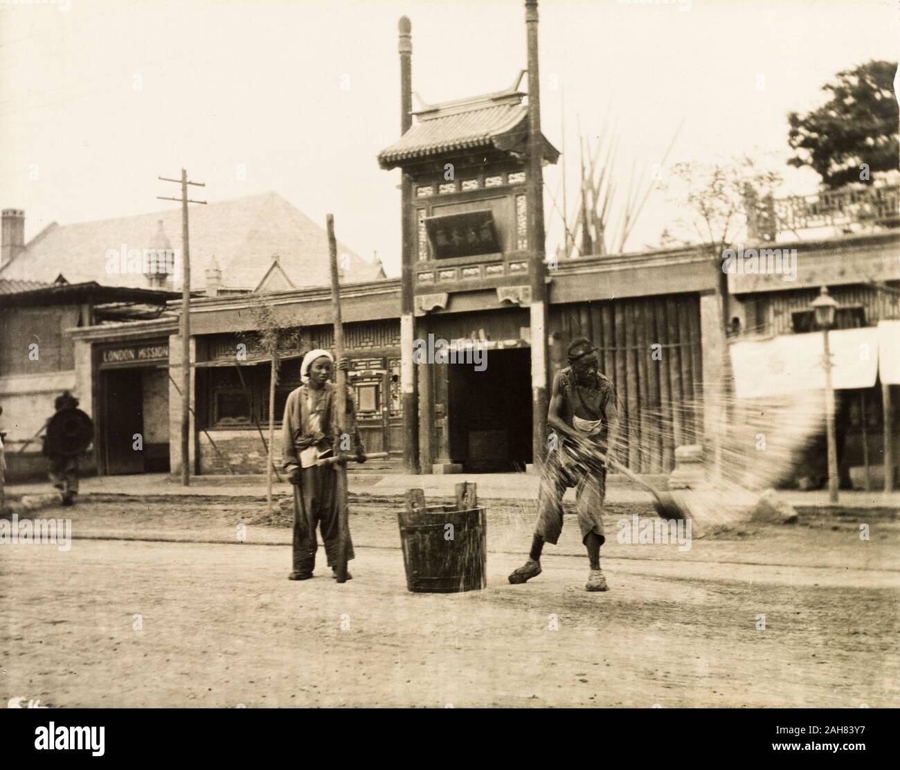 China, A man throws water from a bucket onto the road, probably to control surface dust. Behind him, to the left, is the London Mission building, Beijing.Original manuscript caption: Watering the roadway - London Mission Building to left.The London Mission Building in Beijing was largely destroyed in 1900 so the location of this photo is a mystery as the page to which it is fixed is headed 'Peking', 1909. 1998/028/1/1/139. Stock Photo