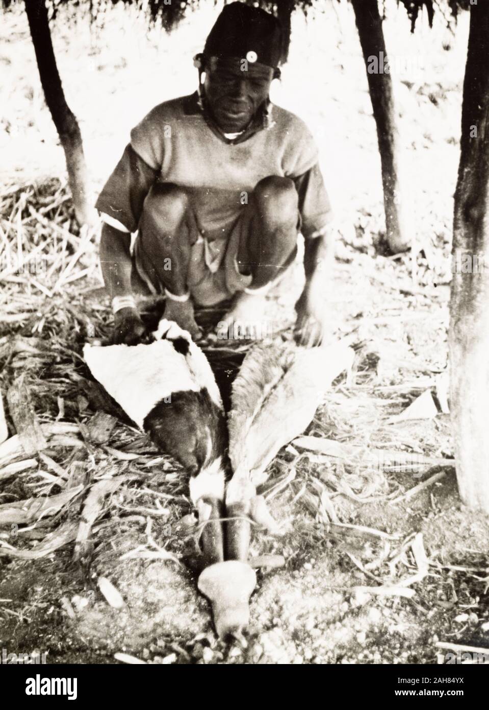 Kenya, A Kikuyu metalsmith operates a pair of goatskin bellows as he forges tools such as knives, pangas and swords in his workshop. Original manuscript caption: A Kikuyu smith. The bellows are goatskins (legs). Knifes, pangas, swords & fire chains are made with these primitive tools. South Nyeri 1936, 1937. 1995/076/1/2/5/20. Stock Photo