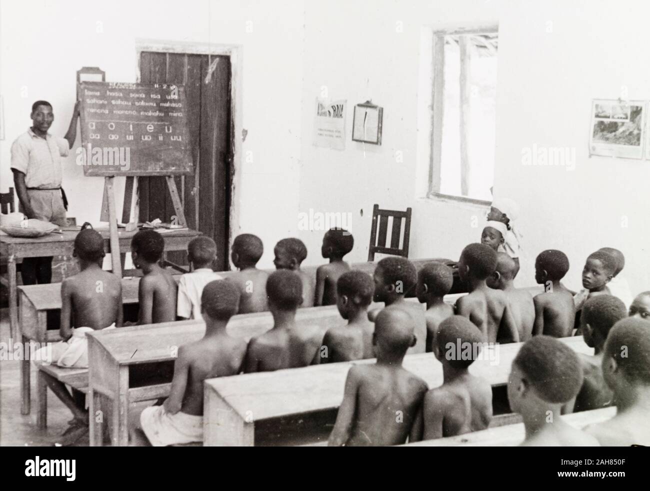 Kenya, Schoolchildren attend a reading lesson at a school in Kenya. Their teacher stands beside a blackboard inscribed with vowel letters and their associated phonetics, perhaps an indication that this is an English lesson. Original manuscript caption: A school at the Coast, 11 February 1947. 1995/076/1/2/7/31. Stock Photo