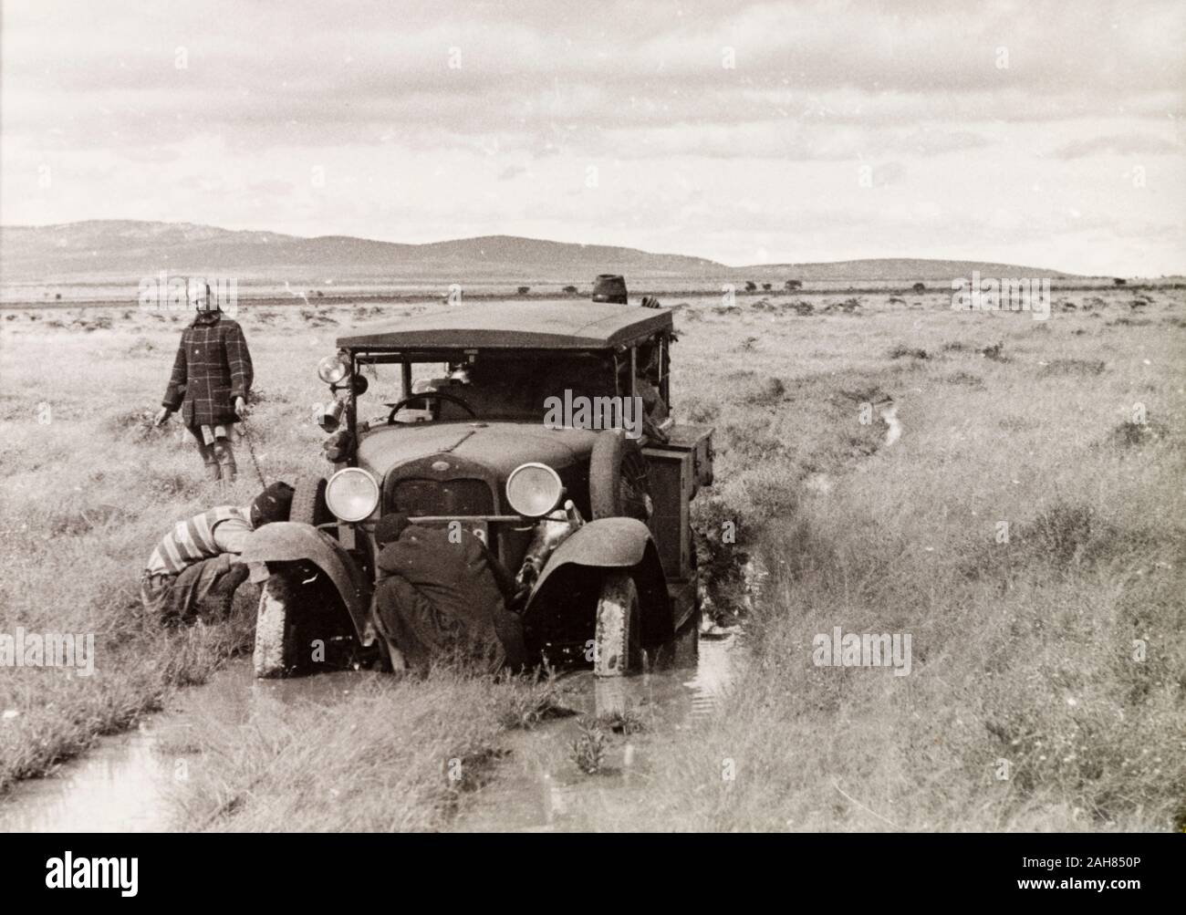 TanzaniaTanganyika, Several men attend to a car, which is stuck in the mud on a flooded country track. This photograph is believed to have been taken on one of the safaris which EH took with the Lindstrom family, most likely the Serengeti safari in 1936 during which they were plagued with bad weather and floods. Original manuscript caption: Motoring in the rains in Northern Tanganyika. Fish on left 1933/34, 1933-1934. 1995/076/1/2/6/27. Stock Photo