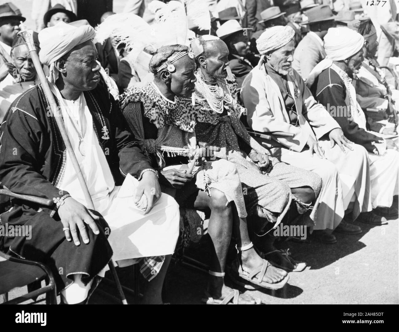 Kenya, Photograph depicting five elderly chiefs sitting at the front of a spectator crowd awaiting the arrival of Princess Elizabeth and the Duke of Edinburgh. The man sitting closest to camera wears a head scarf and holds a staff bearing the Royal insignia. Two other men also wear this head dress. The men sitting in the middle of the front row wear feathered plumes and leopard skin neck ruffs, February 1952. 2001/090/1/4/1/6. Stock Photo