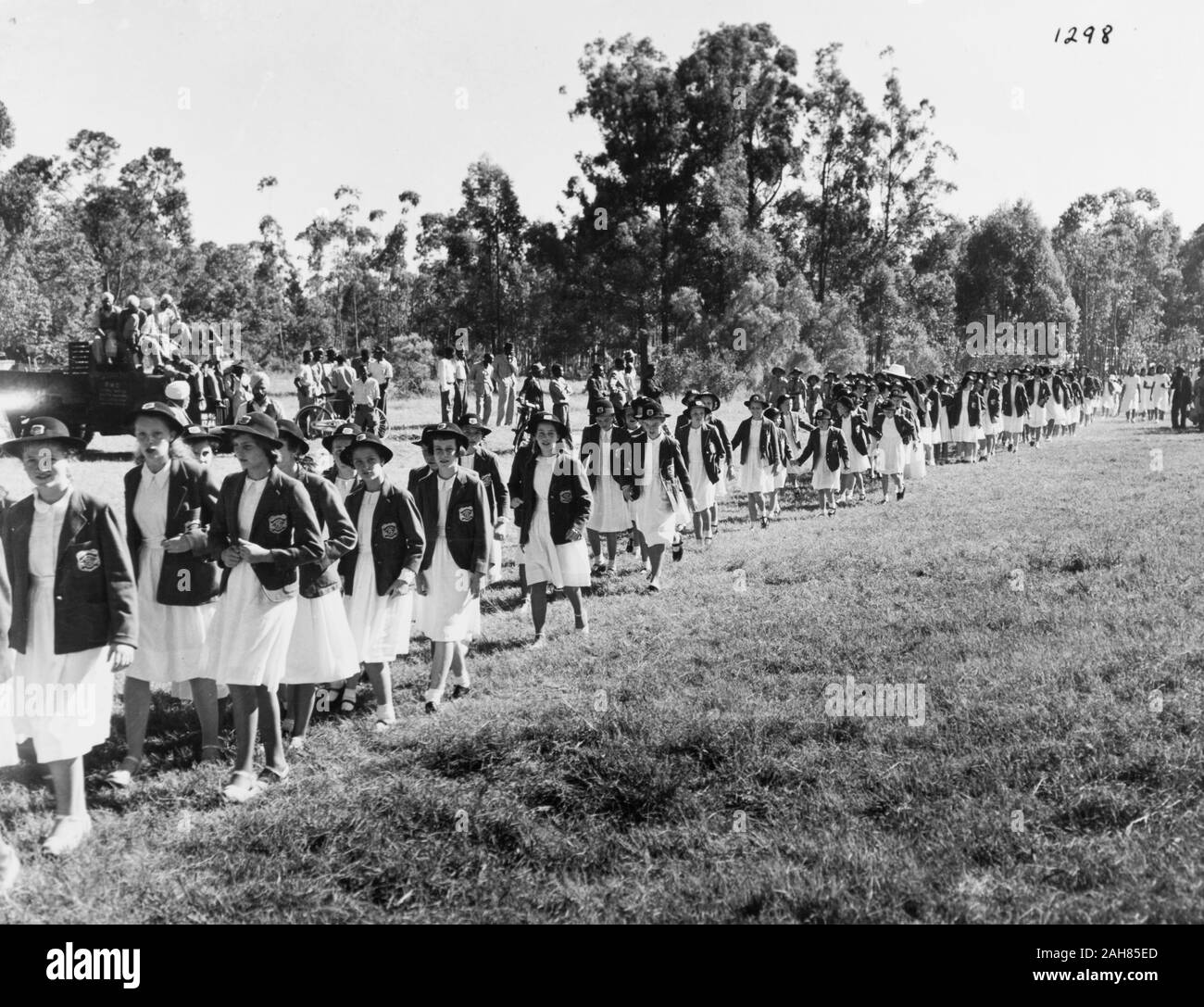 Kenya, A large line of European school girls walk through a field with trees in the background. The girls all wear white dresses with dark blazers and round hats. On the left flank of the line march a small group of African school girls wearing white dresses. In the background of the picture are a group of onlookers; some stand and others sit on a truck, February 1952. 2001/090/1/4/1/29. Stock Photo