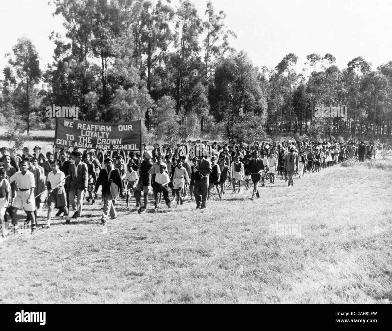 Kenya, A group of Asian teenagers, dressed in school uniform, are escorted by adults as they march through a field with trees in the background. Many of the congregation are carrying Union Jack flags. They are carrying a banner which reads 'We Reaffirm Our Loyalty to the Crown Govt. Asian High School.'', February 1952. 2001/090/1/4/1/27. Stock Photo